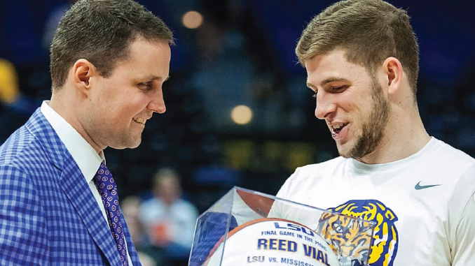 Coach in a blue checkered suit hands a glass trophy to a LSU athlete in a white shirt, both smiling.