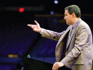 Man in a tan suit delivering a speech at a podium with a microphone in a dark auditorium, gesturing with his right hand.
