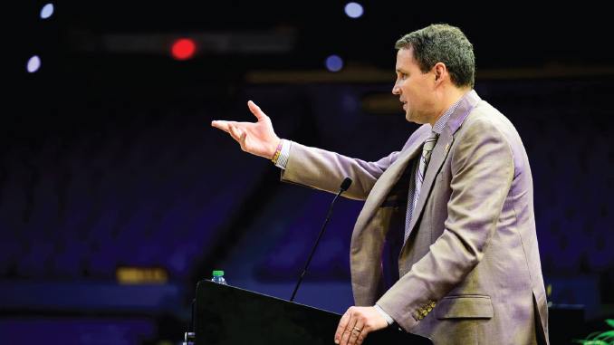Man in a tan suit delivering a speech at a podium with a microphone in a dark auditorium, gesturing with his right hand.