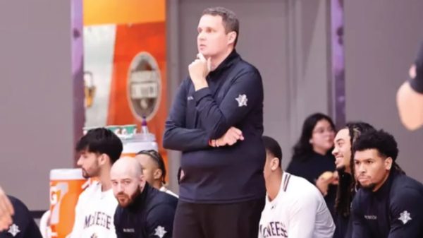 Coach stands with hand on chin while seated players watch during a timeout on the bench.