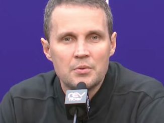 Man speaking at a press conference in front of a purple backdrop, microphone in front.
