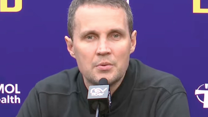 Man speaking at a press conference in front of a purple backdrop, microphone in front.
