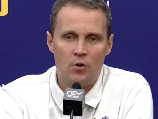 Man in a white polo shirt speaks into a microphone at a press conference with a blue backdrop and logos behind him.