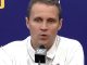 Man in a white polo shirt speaks into a microphone at a press conference with a blue backdrop and logos behind him.