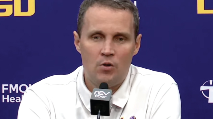 Man in a white polo shirt speaks into a microphone at a press conference with a blue backdrop and logos behind him.