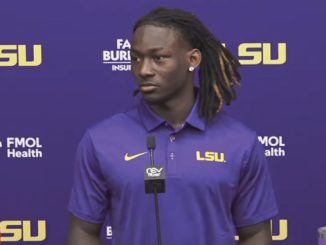 Portrait of a young Black man with dreadlocks wearing a purple LSU polo standing at a press conference microphone, with LSU logos and sponsor banners in the background.
