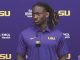 Portrait of a young Black man with dreadlocks wearing a purple LSU polo standing at a press conference microphone, with LSU logos and sponsor banners in the background.