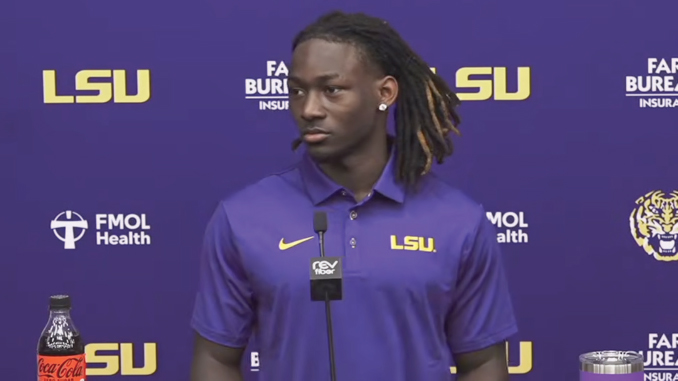 Portrait of a young Black man with dreadlocks wearing a purple LSU polo standing at a press conference microphone, with LSU logos and sponsor banners in the background.
