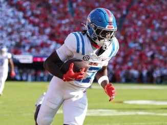 Football player in a white and blue uniform with red gloves runs down the field carrying the ball during a game with a crowded stadium behind him.