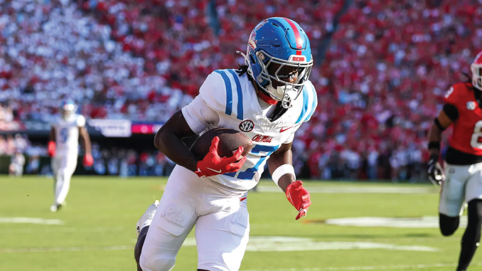 Football player in a white and blue uniform with red gloves runs down the field carrying the ball during a game with a crowded stadium behind him.