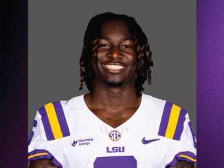 Smiling LSU football player in a white jersey with purple and gold stripes against a gray backdrop.