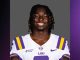 Smiling LSU football player in a white jersey with purple and gold stripes against a gray backdrop.