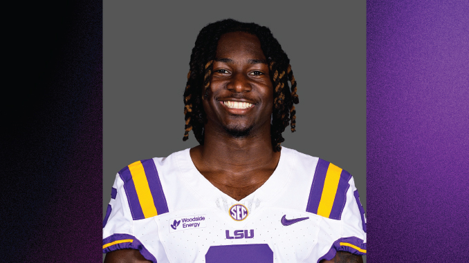 Smiling LSU football player in a white jersey with purple and gold stripes against a gray backdrop.