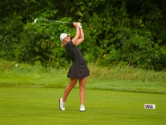 Female golfer in a black outfit swinging a golf club on a green fairway with a USGA marker nearby.