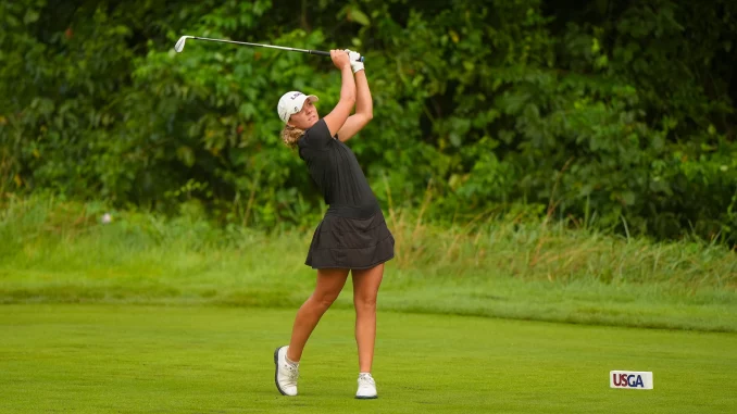 Female golfer in a black outfit swinging a golf club on a green fairway with a USGA marker nearby.