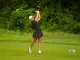 Female golfer in a black outfit swinging a golf club on a green fairway with a USGA marker nearby.