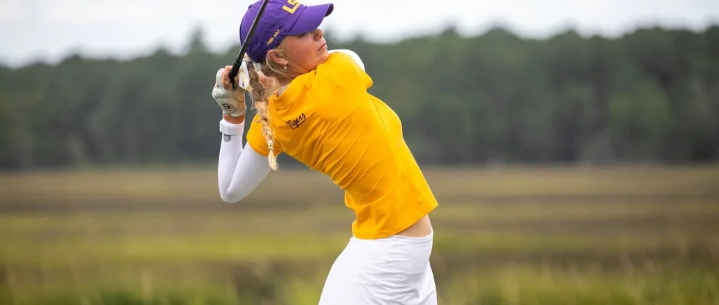 Woman in a yellow polo and white skirt mid-swing with a golf club on a grassy course, wearing a purple cap.