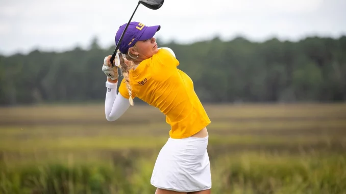 Woman in a yellow polo and white skirt mid-swing with a golf club on a grassy course, wearing a purple cap.