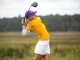 Woman in a yellow polo and white skirt mid-swing with a golf club on a grassy course, wearing a purple cap.