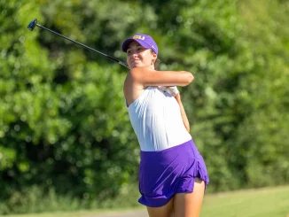 Female golfer mid-swing on a sunny golf course, wearing a purple cap, white top, and purple shorts/skirt with a driver raised behind her.