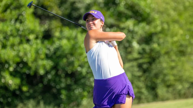 Female golfer mid-swing on a sunny golf course, wearing a purple cap, white top, and purple shorts/skirt with a driver raised behind her.