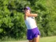 Female golfer mid-swing on a sunny golf course, wearing a purple cap, white top, and purple shorts/skirt with a driver raised behind her.