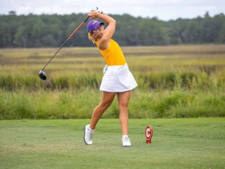 Female golfer in a yellow top and white skirt mid-swing with a driver on a grassy golf course; purple cap and red tee marker nearby.