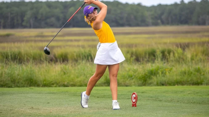Female golfer in a yellow top and white skirt mid-swing with a driver on a grassy golf course; purple cap and red tee marker nearby.