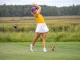 Female golfer in a yellow top and white skirt mid-swing with a driver on a grassy golf course; purple cap and red tee marker nearby.