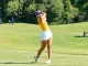 Woman golfer in yellow top and white skirt mid-swing on a green golf course with a cart in the background.