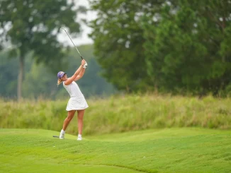 Female golfer in a white outfit mid-swing on a green golf course with trees in the background.