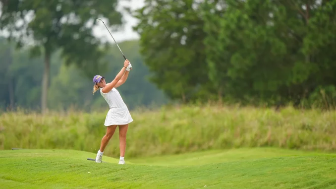 Female golfer in a white outfit mid-swing on a green golf course with trees in the background.