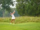 Female golfer in a white outfit mid-swing on a green golf course with trees in the background.