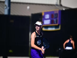 Female tennis player wearing a white visor and black tank top, gripping a racket on a sunlit court with a blurred scoreboard in the background.