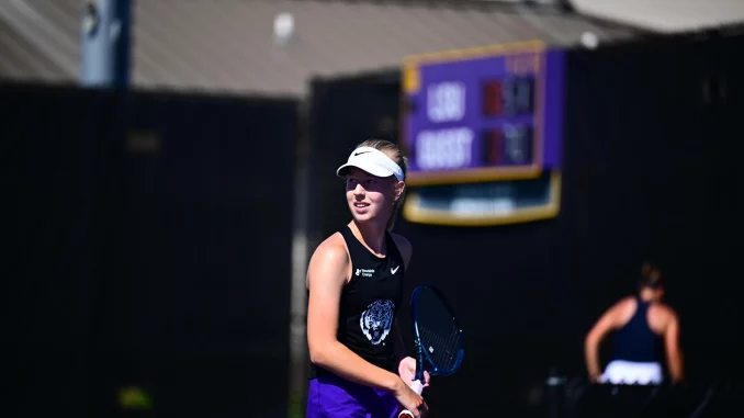 Female tennis player wearing a white visor and black tank top, gripping a racket on a sunlit court with a blurred scoreboard in the background.