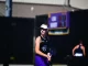 Female tennis player wearing a white visor and black tank top, gripping a racket on a sunlit court with a blurred scoreboard in the background.