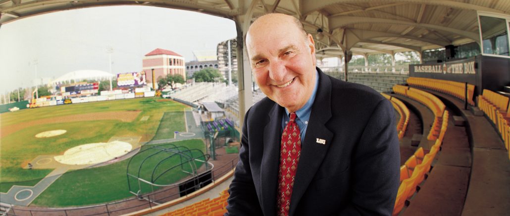 Smiling man in a dark suit and red tie leaning toward the camera in an empty baseball stadium.