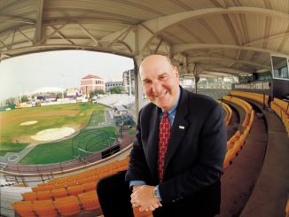 Smiling man in a dark suit and red tie leaning toward the camera in an empty baseball stadium.