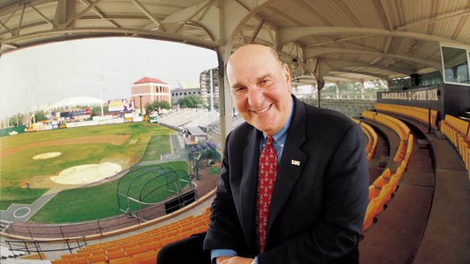 Smiling man in a dark suit and red tie leaning toward the camera in an empty baseball stadium.