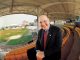 Smiling man in a dark suit and red tie leaning toward the camera in an empty baseball stadium.