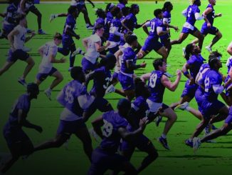 A large group of rugby players in purple uniforms sprint across a bright green field during a match while others chase behind.
