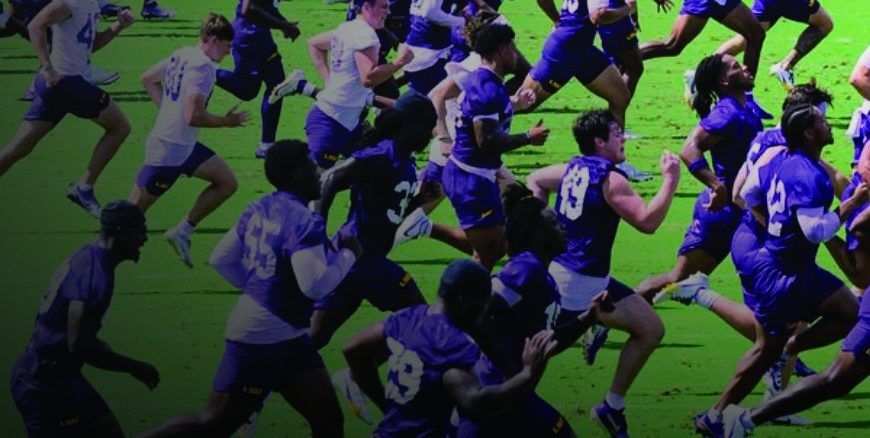 A large group of rugby players in purple uniforms sprint across a bright green field during a match while others chase behind.