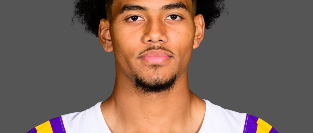 Portrait of a young man with curly black hair wearing an LSU football jersey (white with purple and gold stripes) against a gray background.