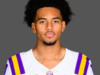 Portrait of a young man with curly black hair wearing an LSU football jersey (white with purple and gold stripes) against a gray background.