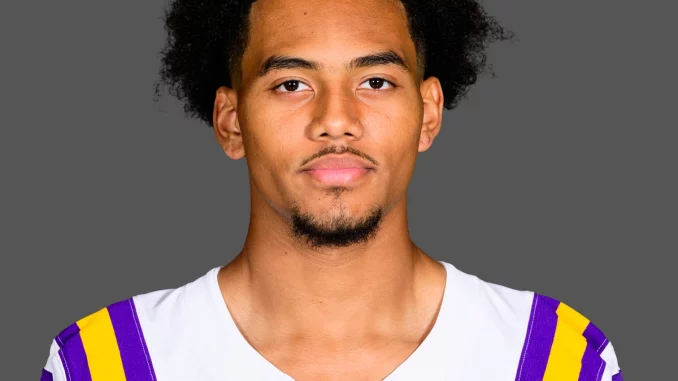 Portrait of a young man with curly black hair wearing an LSU football jersey (white with purple and gold stripes) against a gray background.