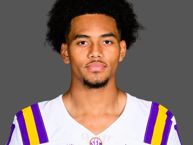 Portrait of a young man with curly black hair wearing an LSU football jersey (white with purple and gold stripes) against a gray background.