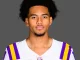 Portrait of a young man with curly black hair wearing an LSU football jersey (white with purple and gold stripes) against a gray background.